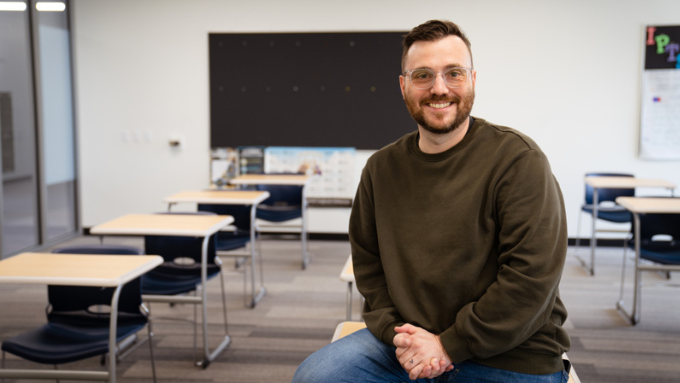 white male with brown hair, glasses and brown crewneck sweater, is perched on a desk in front of an empty classroom
