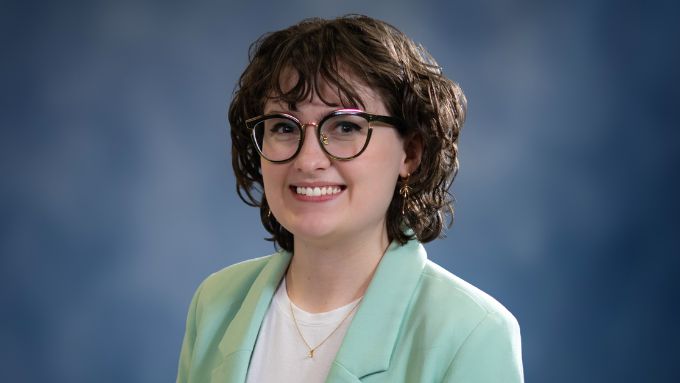 White female with shorter dark curly hair, glasses, white blouse and light green jacket