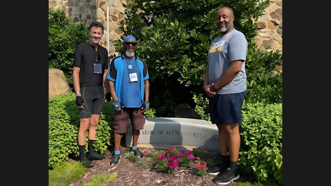 Three men wearing bike gear standing by the Kevin Rice Memorial