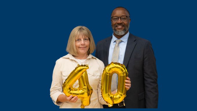 Sharla, an older white woman with shoulder length blonde hair, holds two gold balloons for the number 40. Dr. Spearman, a tall African-American man with black rimmed glasses and a salt and pepper beard stands by her side