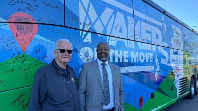 older white male with sunglses and navy jacket stands with African American man wearing a suit; both are in front of a blue bus