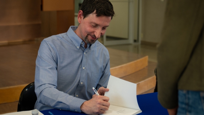 white man with short dark brown hair wearing a light blue long-sleeved button up shirt signs a book