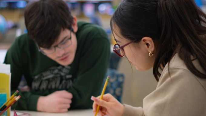 A male student with dark hair glasses wearing a green shirt seated next to a female student with dark and glasses wearing a tan shirt. She is also holding a pencil.