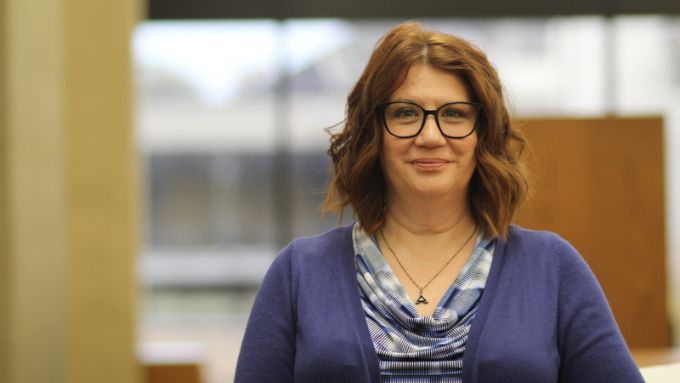 a white woman with dark framed glasses, shoulder length brown hair and an indigo sweater