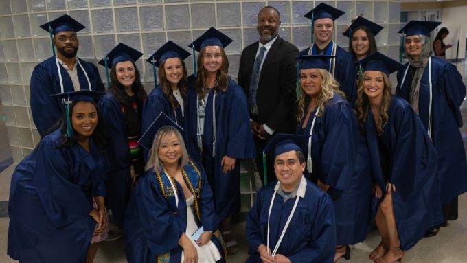 12 graduates in blue caps and gowns stand with the college president