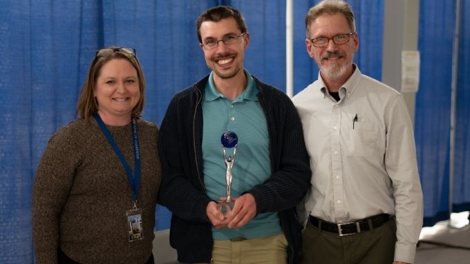 Group of three people, woman on the left and two men on the right. Man in the middle is holding an award.