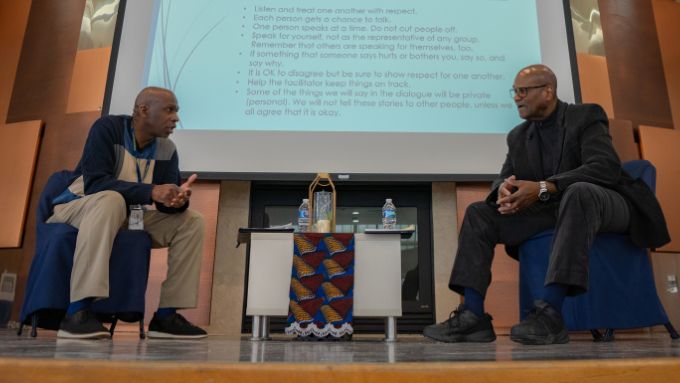 Two African American men seated on a stage with a projector screen in the background.