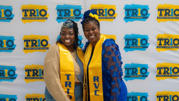 Two African American women stand and smile in front of a backdrop that says TRIO